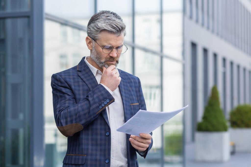Mature gray haired businessman thinking and reading financial document report, senior man in business suit outside office building with bills.